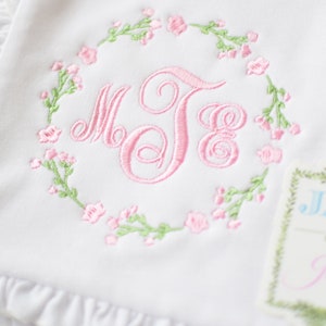 May include: White baby blanket with pink and green embroidered floral wreath and monogram "MJE". A small tag with the text "JAX & Jane" is attached to the blanket.