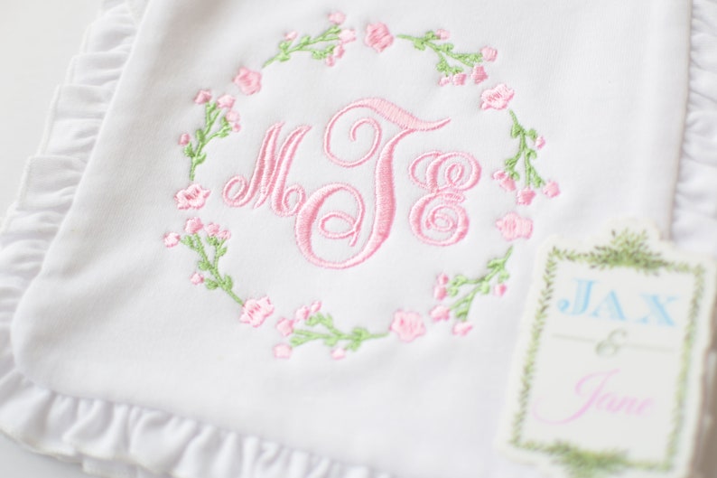 May include: White baby blanket with pink and green embroidered floral wreath and monogram "MJE". A small tag with the text "JAX & Jane" is attached to the blanket.