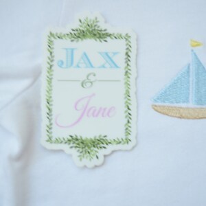 May include: White fabric with a green leafy border frame with the text "Jax & Jane" in blue and pink lettering. A blue sailboat with a yellow sail is embroidered on the fabric.