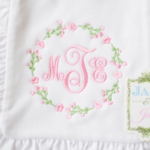 May include: White baby blanket with pink and green embroidered floral wreath and monogram "MJB". A small tag with the text "JAX & Jane" is attached to the blanket.
