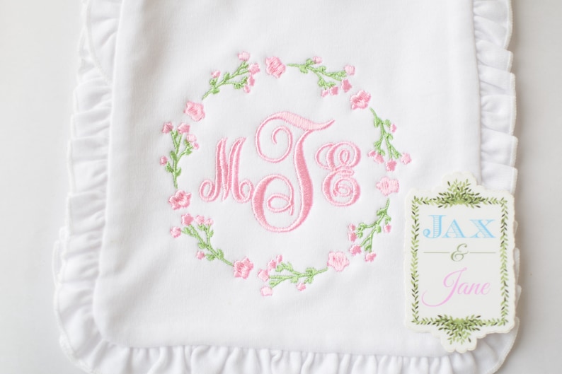 May include: White baby blanket with pink and green embroidered floral wreath and monogram "MJB". A small tag with the text "JAX & Jane" is attached to the blanket.