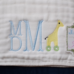 May include: A white baby blanket with blue embroidered letters "MMD" and a yellow embroidered giraffe with green wheels. A white tag with green leaves and the text "Jax & Jane" is in the corner.
