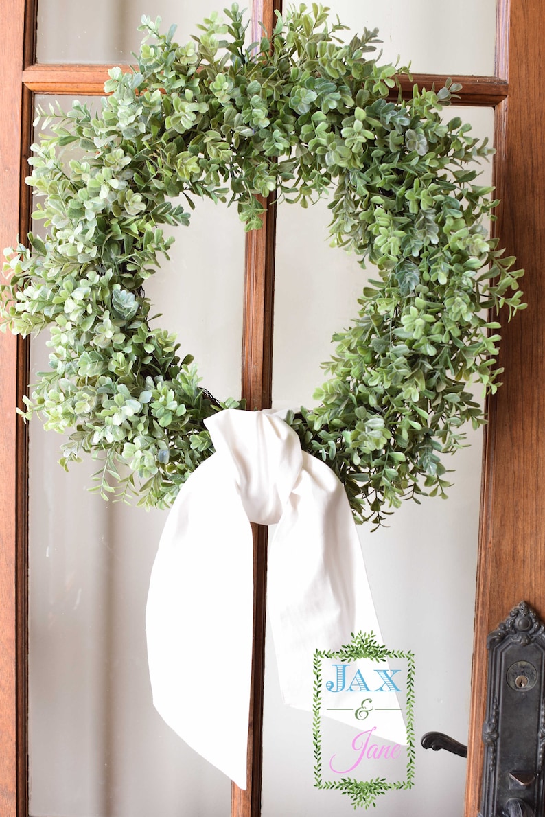 May include: A green leafy wreath with a white fabric bow hanging on a wooden door with a glass pane. The wreath has the text "Jax & Jane" in a green leafy frame.