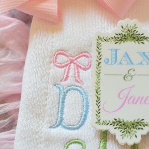 May include: A white embroidered baby onesie with blue and pink bows and the text "Jax & Jane" on a green and white tag.