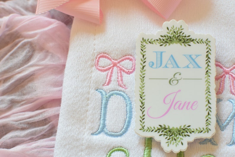 May include: A white embroidered baby onesie with blue and pink bows and the text "Jax & Jane" on a green and white tag.
