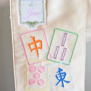 May include: Cream-colored fabric with embroidered Mahjong tiles and a decorative label that reads "Jax & Jane". The tiles feature Chinese characters and symbols in orange, purple, pink, and blue, outlined in various colors.