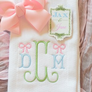 May include: White fabric with green, blue, and pink embroidered letters "DLM" with a pink bow above each letter. A pink bow is on top of the fabric. A sticker with the text "Jax & Jane" is on the fabric.