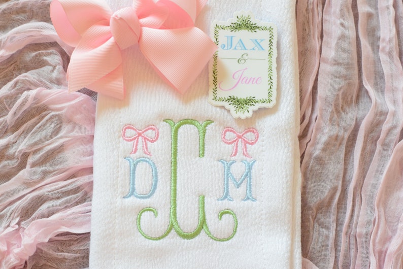 May include: White fabric with green, blue, and pink embroidered letters "DLM" with a pink bow above each letter. A pink bow is on top of the fabric. A sticker with the text "Jax & Jane" is on the fabric.