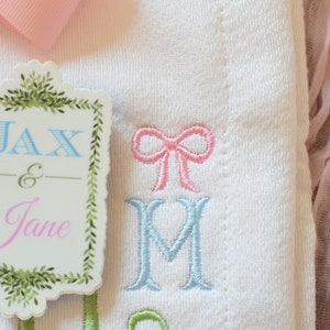 May include: A white embroidered baby blanket with a pink bow and the names Jax & Jane on a green and white tag.