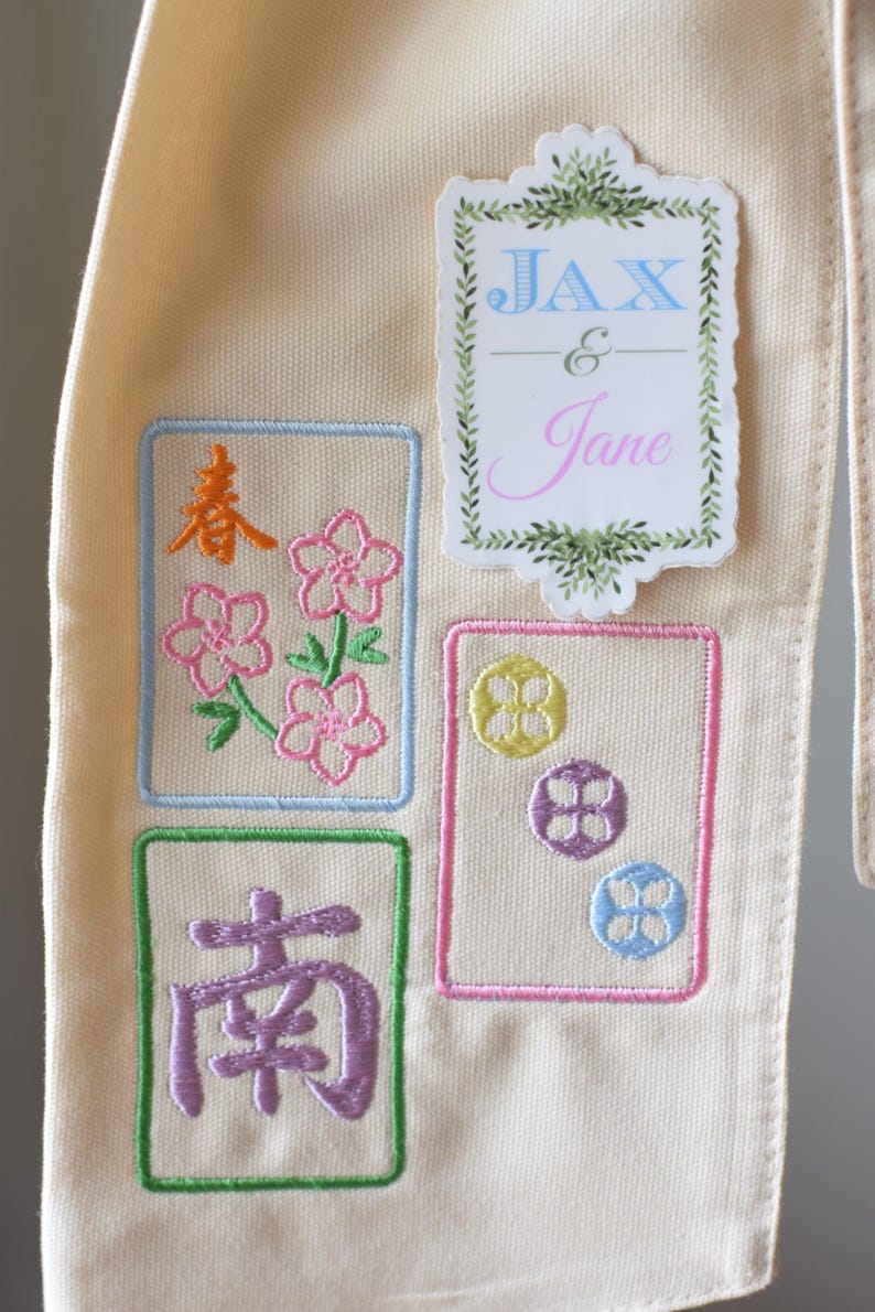 May include: Cream-colored fabric with embroidered patches. Patches include floral designs, Chinese characters, and the names "Jax & Jane". The embroidery features pink, blue, yellow, and purple colors.