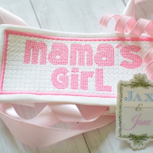May include: White fabric with pink embroidered lettering that reads "mama's girl". A small white tag with green foliage border and blue and pink lettering reads "Jax & Jane".
