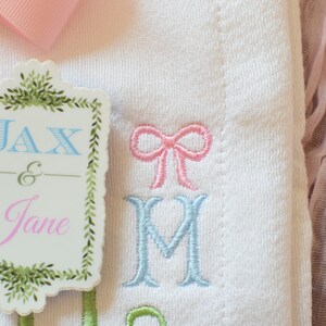 May include: A white embroidered baby blanket with a pink bow and the names Jax & Jane on a green and white tag.
