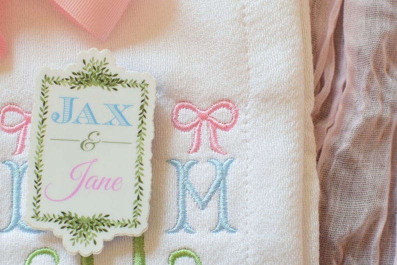 May include: A white embroidered baby blanket with a pink bow and the names Jax & Jane on a green and white tag.