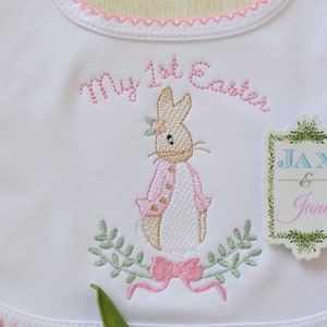 Könnte beinhalten: Weißer gestickter Baby-Lätzchen mit rosa Besatz. Das Lätzchen zeigt ein rosa und braunes Kaninchen mit einer rosa Schleife, grünen Blättern und dem Text "My 1st Easter". Ein separates Etikett mit grünen Blättern und dem Text "Jax & Jane" ist am Lätzchen befestigt.