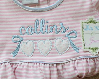 May include: Pink and white striped baby dress with blue embroidered hearts and the name "collins" on the front. A tag with the text "JAX & Jane" is attached to the dress.