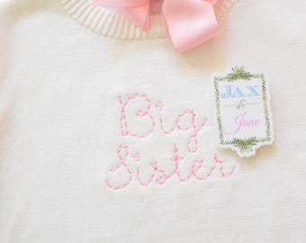 May include: White sweater with pink embroidery that reads "Big Sister". A small white tag with green foliage border reads "Jax & Jane" in blue and pink lettering.