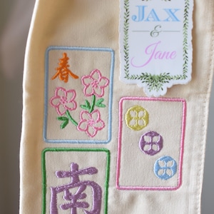 May include: Cream-colored fabric with embroidered designs. Features four rectangular embroidered patches with floral and Chinese character motifs in various colors. A sticker with the names "Jax & Jane" is also present.