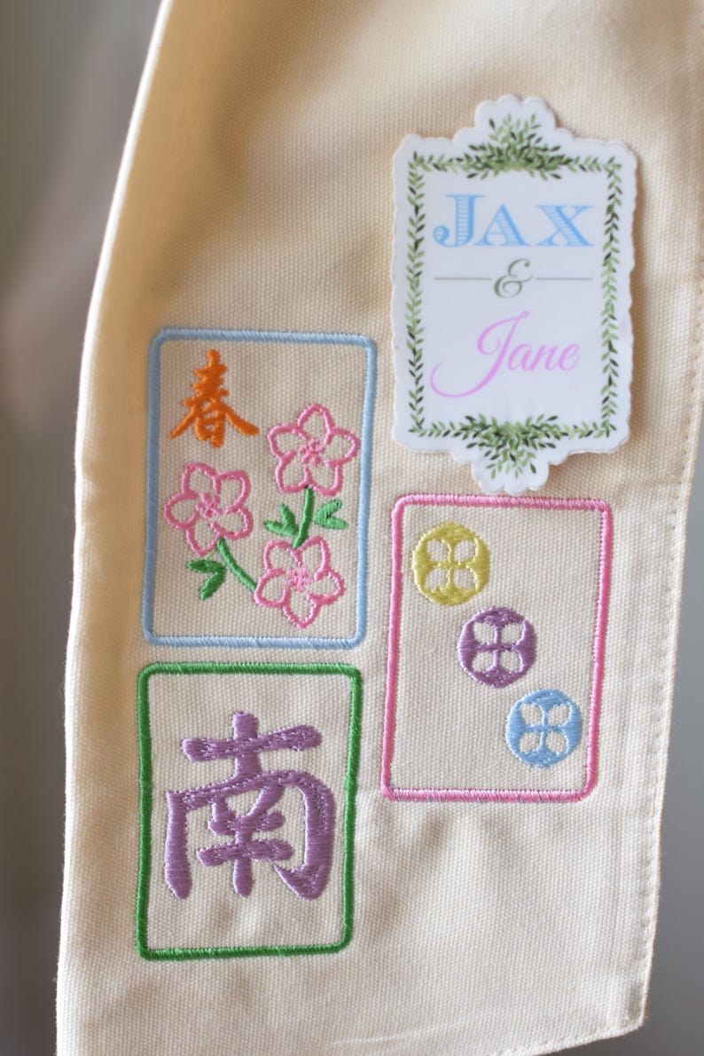 May include: Cream-colored fabric with embroidered designs. Features four rectangular embroidered patches with floral and Chinese character motifs in various colors. A sticker with the names "Jax & Jane" is also present.