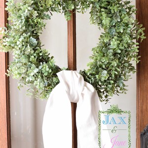 May include: A green leafy wreath with a white fabric bow tied around it. The wreath is hanging on a wooden door with a glass pane. The text "JAX & Jane" is written on a sign in the bottom right corner of the image.