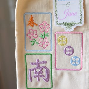 May include: Cream-colored fabric with embroidered designs. Features floral and geometric patterns in pink, orange, green, and blue. Includes the text "JAX & Jane" in a decorative frame.