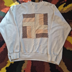 May include: A cream-colored crewneck sweatshirt featuring a square patch on the chest. The patch is constructed from rectangular fabric pieces in alternating floral brown and solid light peach patterns with visible raw, frayed edges.