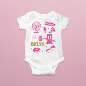 May include: White baby onesie with a pink graphic design featuring the Brooklyn Bridge, a Ferris wheel, a hot dog, a donut, a slice of pizza, and an ice cream cone. The text "BKLYN" and "BROOKLYN, NY" are also included in the design.
