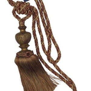 May include: A brown and gold tassel tieback with a decorative ball and a braided cord. The cord is a combination of brown, gold, and cream colors. The label reads "Trims & Traditions Tassel"