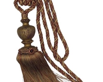 May include: A brown and gold tassel tieback with a decorative ball and a braided cord. The cord is a combination of brown, gold, and cream colors. The label reads "Trims & Traditions Tassel"