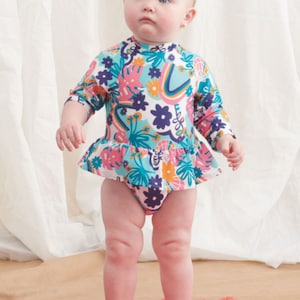 Puede incluir: Un bebé con un traje de baño de manga larga blanco, azul y rosa con estampado floral. El traje de baño tiene un volante en la parte inferior y un cuello alto.