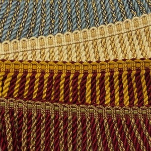 May include: Close-up of decorative trim in various colors. The trim includes blue, beige, gold, and burgundy. The trim has a braided rope design with a fringe. The trim is likely used for upholstery or drapery.