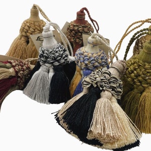 Könnte beinhalten: Sortiment von sechs dekorativen Quasten in verschiedenen Farben und Texturen, darunter Beige, Grün, Schwarz, Weiß und Rot. Die Quasten sind aus gewebtem Stoff und haben einen Fransen unten.