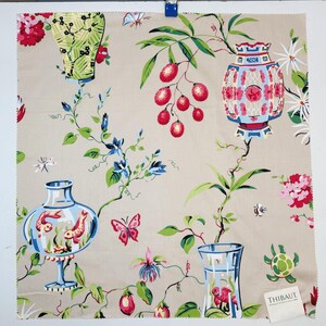 Può includere: Un campione di tessuto beige con un colorato motivo botanico e decorativo. Il design include litchi rossi, lanterne blu e rosa, vasi verdi con pesci, farfalle e piccole tartarughe su uno sfondo marrone chiaro con il testo THIBAUT.