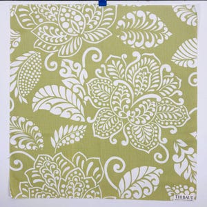 Könnte beinhalten: Ein quadratisches Stoffstück mit hellolivgrünem Hintergrund und einem sich wiederholenden weißen Blumen- und Blattmuster. Das Design zeigt große stilisierte Blüten und Blätter mit feinen Details. Ein kleines weißes Etikett trägt die Aufschrift THIBAUT.