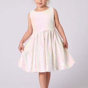 May include: A sleeveless, knee-length dress covered in iridescent sequins. The dress has a fitted bodice and a flared skirt. The girl is wearing gold-colored shoes with bows. The background is plain white.