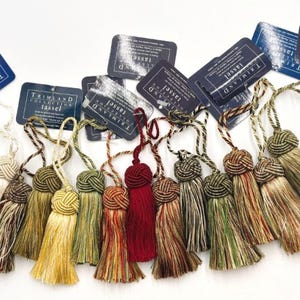 Może przedstawiać: Kolekcja ozdobnych chwostów w różnych kolorach, w tym złotym, zielonym i czerwonym. Każdy chwost ma węzeł na górze i długi, postrzępiony dół. Etykiety głoszą "TRIMLAND COLLECTION TASSEL".