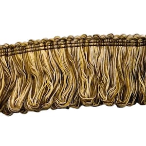 May include: Close-up of decorative fringe trim. The trim features a woven top in shades of brown and gold, with long, flowing fringe in similar colors. The fringe is made of multiple strands of yarn, creating a textured effect.