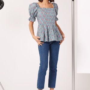 May include: A blue and white floral print blouse with puffed sleeves and a square neckline. The blouse has a fitted bodice and a flowy peplum hem. The model is wearing blue jeans and tan heeled sandals.