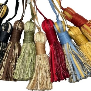 Puede incluir: Una colección de borlas decorativas en varios colores, incluyendo negro, marrón, verde, rojo, azul y dorado. Cada borla presenta una parte superior envuelta y una parte inferior larga y con flecos, ideal para la decoración del hogar o proyectos de manualidades.