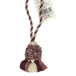 以下が含まれることがあります： ピンク、白、茶色の房飾りで、編み紐が付いています。房飾りは撚り糸で作られており、ふわふわとした質感です。紐は3本の糸でできており、それぞれ色が異なります。紐に付いているタグには、「CORD 61% POLY 19%CO 1%A 3%WD 91962 08812 0 MATERIALS MADE IN USA RN 23003 ASSEMBLED IN MEX」と書いてあります。