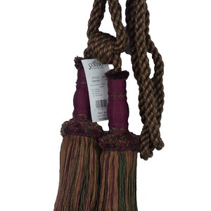 May include: A pair of burgundy and green tassel tiebacks with a rope cord. The tassels are made of a woven fabric with a fringe. The rope cord is twisted and has a brown and green color scheme. The tiebacks are labeled with the brand name "COISO International" and the style number "022045".