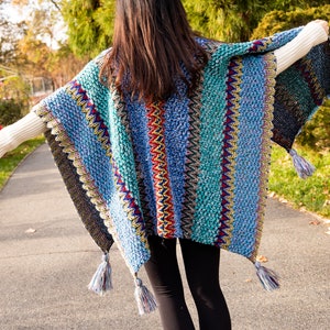 Puede incluir: Un poncho de crochet a rayas de colores con flecos. El poncho es azul, verde, rojo, amarillo y negro.