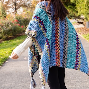 Puede incluir: Un poncho de punto azul y turquesa con rayas de colores y borlas. El poncho est&aacute; hecho de un hilo suave y peludo.