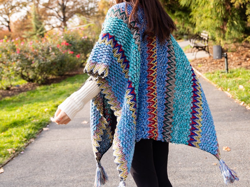 Puede incluir: Un poncho de punto azul y turquesa con rayas de colores y borlas. El poncho est&aacute; hecho de un hilo suave y peludo.
