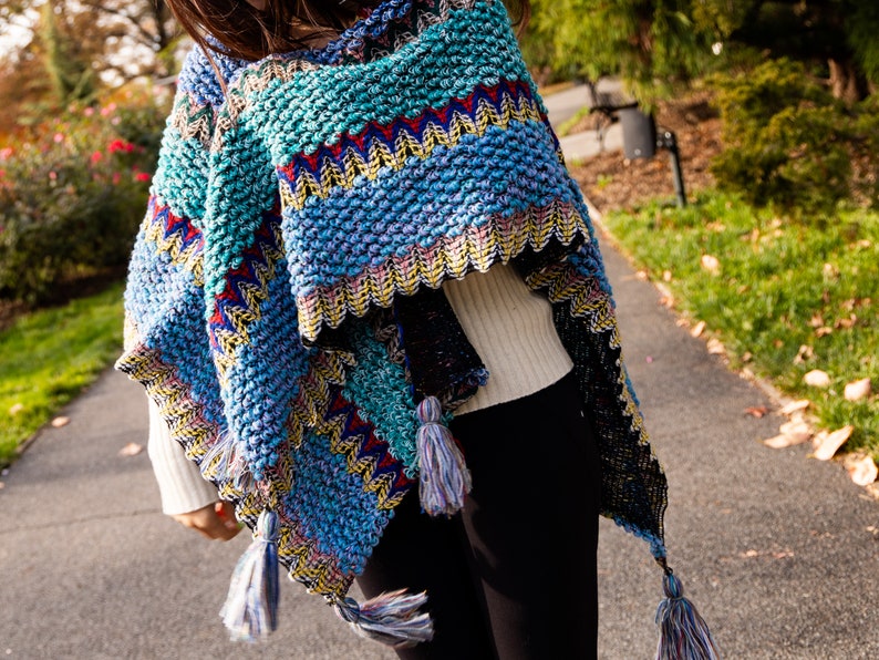 Puede incluir: Un poncho de punto colorido con flecos. El poncho est&aacute; hecho de un hilo suave y texturizado y presenta una variedad de colores, incluyendo azul, verde, rojo y amarillo.
