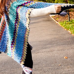 Puede incluir: Una mujer lleva un poncho de punto de colores con flecos. El poncho tiene un patr&oacute;n azul, verde, amarillo, rojo y blanco.