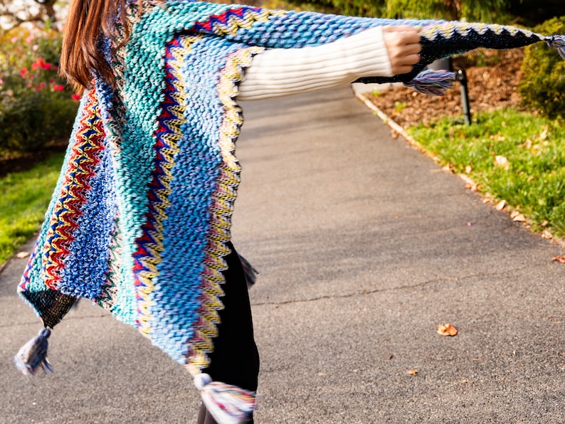 Puede incluir: Una mujer lleva un poncho de punto de colores con flecos. El poncho tiene un patr&oacute;n azul, verde, amarillo, rojo y blanco.