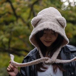 May include: A woman wearing a white fuzzy hooded hat with bear ears. The hat is tied at the neck with a long scarf. She is wearing a black leather jacket and a white shirt.
