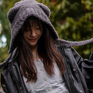 May include: A woman wearing a gray fuzzy hooded hat with bear ears and a black leather jacket. The hat is tied around her neck and she is looking down.