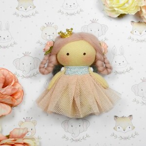 Puede incluir: Muñeca hecha a mano con un vestido rosa claro y una corona dorada. La muñeca tiene cabello castaño claro y está adornada con flores rosas. El fondo presenta un patrón de animales, incluyendo elefantes, conejos y zorros.