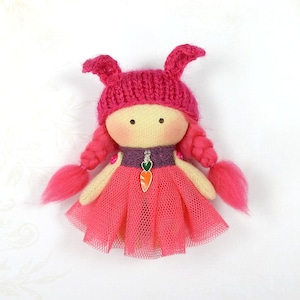Puede incluir: Una muñeca de tela rosa con pelo de lana rosa, que lleva un gorro de punto rosa con orejas de conejo, un corpiño morado y una falda de tul rosa. La muñeca tiene un amuleto de zanahoria alrededor del cuello.